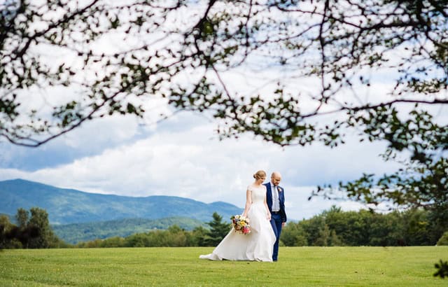 Bride and groom walking on lush green field with mountain view in Vermont.