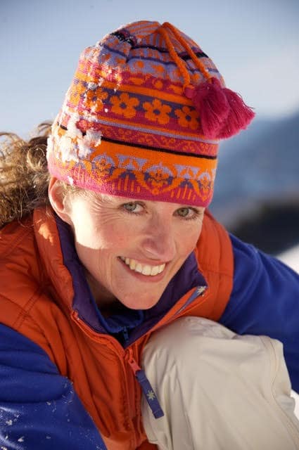 Smiling woman in winter gear with colorful knit hat in snowy landscape.