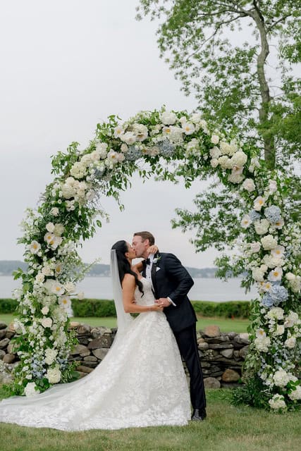 Bride and groom kissing under floral wedding arch by the lake.
