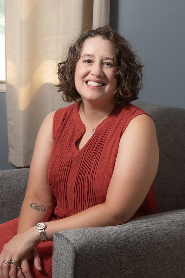 Portrait of a smiling woman in a red dress, sitting on a gray sofa.