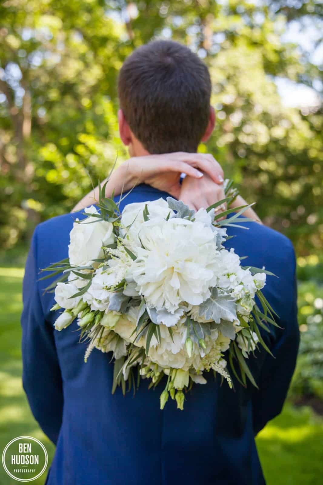 Groom with white floral bouquet on his back outdoors in a garden setting.